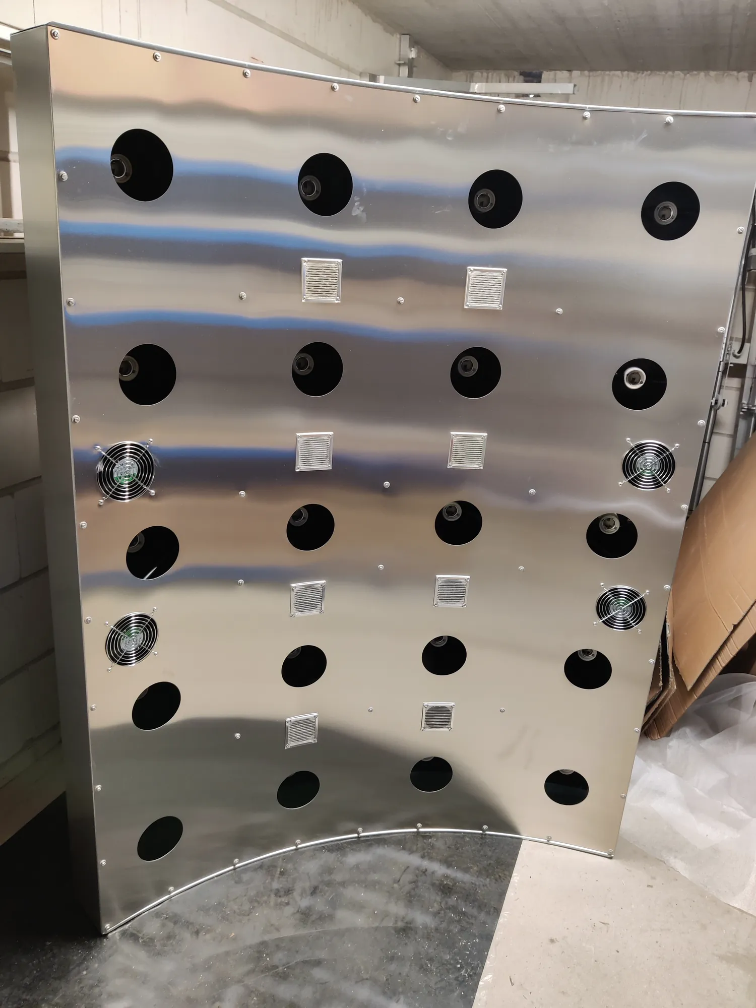 Eine gewölbte Schale aus gebürstetem Edelstahl steht aufrecht auf dem Werkstattboden. In den Bohrungen sitzen schon die Lampenfassungen, dazwischen quadratische Lüftungsgitter und die Lüfter; die Strahler fehlen noch.